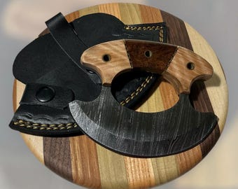 Könnte beinhalten: Ein Damaststahlmesser mit Holzgriff und schwarzer Lederscheide. Der Griff weist helle und dunkle Holzsegmente auf. Das Messer wird auf einem runden Holzschneidebrett mit verschiedenen Holztönen präsentiert.
