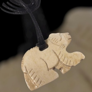 May include: Cream-colored pendant carved in the shape of a bear, suspended from a black cord. The bear pendant features etched details and a smooth finish. A unique, handcrafted necklace.
