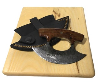 Könnte beinhalten: Ein gebogenes Messer aus Damaststahl mit einem Holzgriff und einer Lederscheide. Das Messer liegt auf einem Holzbrett.