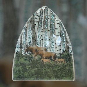 Può includere: Un pezzo decorativo in vetro con una scena di foresta con betulle e una famiglia di alci. L'opera d'arte ha una forma triangolare con la parte superiore arrotondata. Gli alci sono marroni e gli alberi sono bianchi con segni neri.
