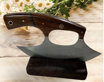 Peut inclure: Un couteau mezzaluna avec un manche en bois foncé et une lame incurvée. Le manche est orné de rivets en laiton et d'un emblème métallique décoratif. Le couteau est présenté sur un socle en bois foncé, soulignant son design unique et son savoir-faire.