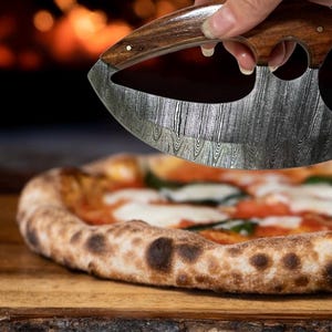 Op de afbeelding: Een pizzasnijder met een gebogen, patroonvormig mes en een houten handvat wordt boven een pizza gehouden. De pizza heeft een goudbruine korst, belegd met kaas, tomaten en basilicum. De achtergrond is wazig.