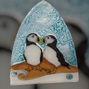 Peut inclure: Une pièce décorative en verre représentant deux macareux moines enlacés. Les oiseaux ont des corps noirs et blancs, des becs jaunes et se tiennent sur une surface marron sur un fond bleu texturé. La pièce a un design artistique unique.