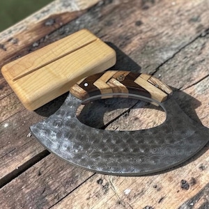 May include: A Damascus steel herb chopper with a wooden handle and a wooden stand. The chopper has a curved blade with a decorative pattern. The handle is made of multiple pieces of wood. The stand is rectangular with a groove.