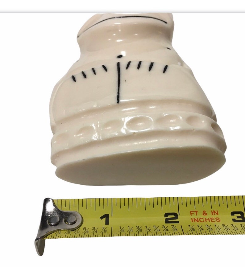 May include: A white ceramic figurine of a person wearing a dress with a black design. The figurine is approximately 2 inches tall.
