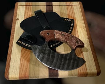 May include: A wooden handled mezzaluna knife with a curved blade rests on a striped wooden cutting board. A black leather sheath with a belt loop is next to the knife. The knife blade is dark with a textured surface.