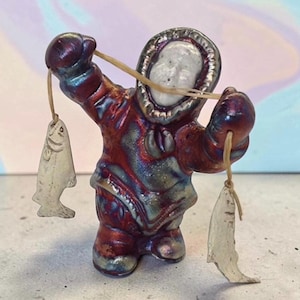 May include: A small ceramic figurine of a person wearing a hooded parka with a metallic red and blue finish. The figure holds a thin string in each hand, with a small, off-white ceramic fish dangling from each end of the string in front of them.
