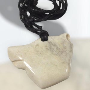 Op de afbeelding: Crèmekleurige hanger in de vorm van een berenkop, hangend aan een zwart koord. De berenhanger heeft gesneden details en een gladde afwerking. Het koord is aan de bovenkant geknoopt, waardoor een lus ontstaat om te dragen.