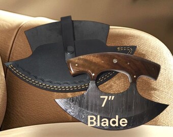 Könnte beinhalten: Ein halbmondförmiges Ulu-Messer mit einem Griff aus dunklem Holz und einer gemusterten Metallklinge, auf der der Text 7" Blade steht. Das Messer wird von einer schwarzen Lederscheide mit gelben Nähten und einem Druckknopfverschluss begleitet.