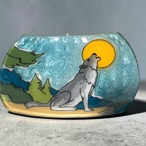 May include: A curved glass decorative piece featuring a gray wolf howling at a large yellow moon. The background depicts a textured light blue sky, green pine trees, and a tan ground. The design is rendered in a flat, illustrative style.