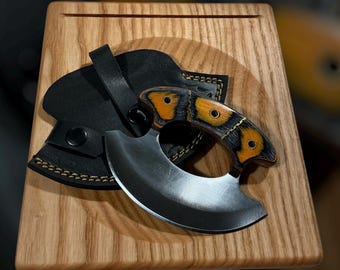 May include: A handcrafted, curved blade knife with a wooden handle featuring orange and black accents. The knife rests on a wooden cutting board, accompanied by a black leather sheath. The blade has a silver finish.
