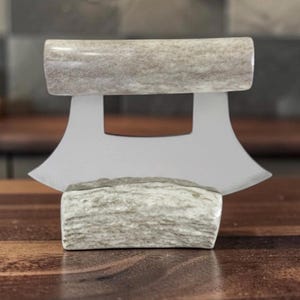 Puede incluir: Un cuchillo de cocina con una hoja de metal plateado curvada y un mango hecho de una pieza de asta natural texturizada de color claro. La hoja presenta un diseño ancho en forma de media luna con un recorte rectangular en el centro.