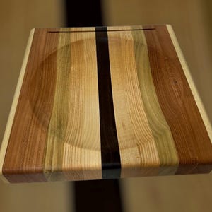 May include: A square wooden cutting board with a recessed center. The board is made of various wood tones, including light, medium, and dark browns, with a black stripe down the center. The edges are rounded.