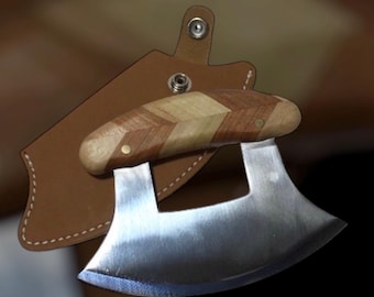 Peut inclure: Un couteau mezzaluna en acier inoxydable avec un manche en bois à chevrons. Le couteau repose sur un étui en cuir marron avec une fermeture à bouton-pression. Le manche est en bois clair et foncé.
