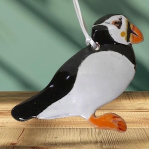 May include: A ceramic ornament shaped like a puffin with black and white markings and an orange beak. The ornament is hanging from a white string.