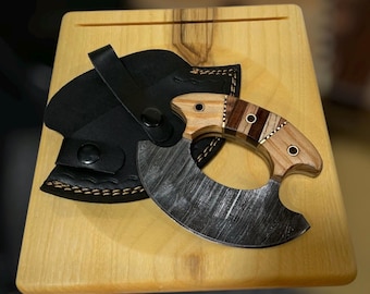 Könnte beinhalten: Ein handgefertigtes Messer mit gebogener Klinge und einem Holzgriff mit einer Mischung aus hellen und dunklen Holztönen. Die Klinge hat eine strukturierte, dunkle Oberfläche. Es liegt auf einem hellen Holzschneidebrett mit einer schwarzen Lederscheide.