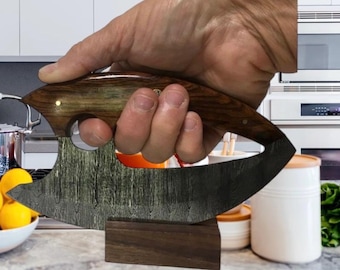 Könnte beinhalten: Ein einzigartiger, gebogener Kräuterhacker mit einer dunklen, gemusterten Klinge und einem Holzgriff. Der Griff hat ein Fingerloch zum Greifen und wird in einer Hand gehalten. Der Hacker ruht auf einem kleinen Holzblock.