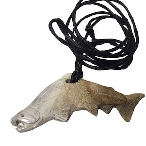 May include: A carved stone pendant in the shape of a fish, suspended from a black cord. The fish is light brown with carved details, including fins, gills, and an open mouth. The cord is knotted at the top.