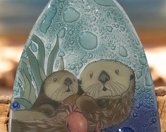 May include: A decorative glass piece featuring two otters and a crab. The otters are brown and tan, with the crab in pink. The background is blue with bubble-like details and seaweed. The piece is triangular in shape.