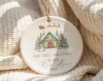 May include: A white ceramic Christmas ornament with a festive illustration of a green house, snowman, and Santa's sleigh. The text reads "OUR FIRST CHRISTMAS IN OUR NEW HOME 2025". A gold string is attached for hanging.