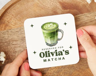 May include: Square white coaster featuring a matcha latte illustration in a glass mug. The coaster reads "RESERVED FOR Olivia's MATCHA" in green. Four small black star shapes are in each corner of the coaster.