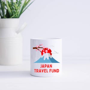 May include: White ceramic mug with a graphic of Mount Fuji, a red sun, cherry blossoms, and the text "JAPAN TRAVEL FUND" in red. The mug is cylindrical and sits on a white surface.