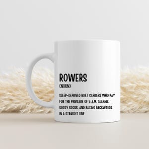 May include: White ceramic mug featuring the word "ROWERS" in black, with a definition below. The definition describes rowers as sleep-deprived boat carriers. The mug has a curved handle and is set against a neutral background.