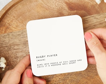 May include: White coaster with black text that reads "RUGBY PLAYER (NOUN) RUNS INTO PEOPLE AT FULL SPEED AND CALLS IT A WEEKEND WELL SPENT"