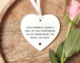May include: A white heart-shaped ornament with a ribbon, featuring the text "YOUR KINDNESS LEAVES A TRAIL OF LIGHT EVERYWHERE YOU GO. NEVER DOUBT THE IMPACT YOU HAVE." A pink rose is next to the ornament.