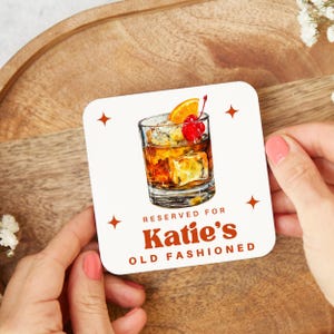 May include: Cream-coloured square coaster featuring an Old Fashioned cocktail illustration, garnished with orange and cherry. Text reads "Reserved for Katie's Old Fashioned". Decorative starbursts are in each corner.