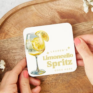 May include: A square coaster with a watercolour illustration of a limoncello spritz cocktail with lemon slices. The coaster has the text "Lauren's Limoncello Spritz Goes Here" in gold lettering.
