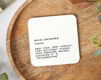 May include: White coaster with black text that reads "WILD SWIMMER (NOUN) PART FISH, PART LUNATIC. THEY WILLINGLY PLUNGE INTO FREEZING WATERS, CLAIMING IT'S 'INVIGORATING'"
