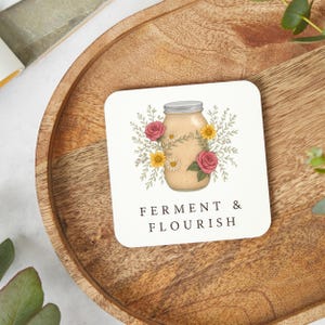 May include: Square coaster featuring a jar illustration. The jar is filled with a beige substance and surrounded by pink roses, yellow daisies, and green leaves. The words "FERMENT & FLOURISH" are printed below the jar in a serif font.
