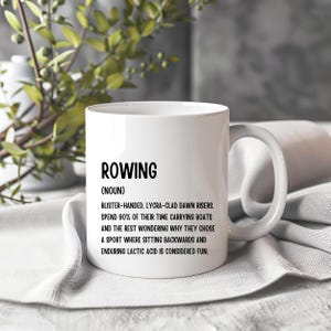 May include: White ceramic mug featuring the word "ROWING" in black, with a humorous definition below. The mug has a curved handle and is placed on a white cloth, with foliage visible in the background.
