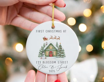 May include: White ceramic Christmas ornament with a watercolor illustration of a house, snowman, and Christmas trees. The text reads "First Christmas At" and "123 Blossom Street Chloe & Jacob 2025". A gold string is attached for hanging.
