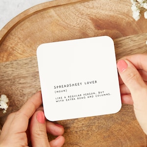 May include: White coaster with black text that reads "Spreadsheet Lover (Noun) Like a regular person, but with extra rows and columns."