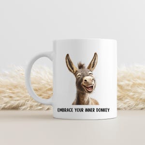 Puede incluir: Taza de cerámica blanca con una ilustración de un burro sonriente. El burro está representado en tonos marrones y grises, con una gran sonrisa y orejas prominentes. El texto "EMBRACE YOUR INNER DONKEY" está impreso debajo. La taza tiene un asa curva.