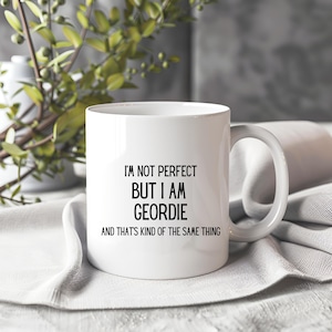 Puede incluir: Taza de cerámica blanca con asa curva. La taza presenta texto negro que dice: "I'M NOT PERFECT BUT I AM GEORDIE AND THAT'S KIND OF THE SAME THING". La taza está sobre un paño blanco.