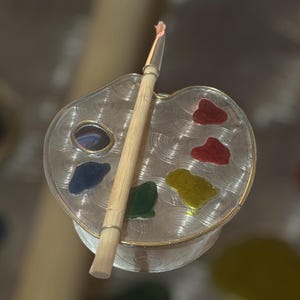 May include: A silver-toned artist's palette-shaped container with a wooden paintbrush. The palette has colorful paint splotches in red, yellow, green, and blue. The paintbrush is positioned across the palette.