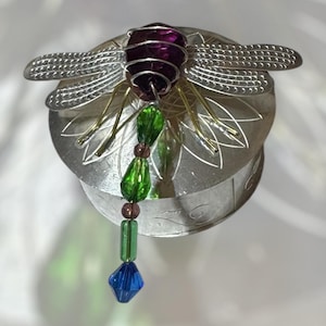 Könnte beinhalten: Eine silberfarbene Metallbox mit einem Libellen-Design. Die Libelle hat einen lila Edelsteinkörper, silberne Flügel und eine Perlenkette aus grünen, lila und blauen Edelsteinen.