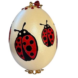 May include: A white egg with a gold stand and a red crystal hanging from the top. The egg is painted with three red and black ladybugs.