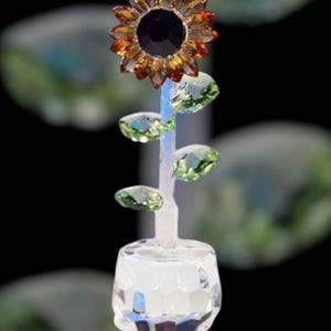 Könnte beinhalten: Eine Kristall-Sonnenblumenfigur mit grünen Blättern und einem klaren Kristallsockel. Die Sonnenblume hat eine dunkelbraune Mitte und orangefarbene Blütenblätter.