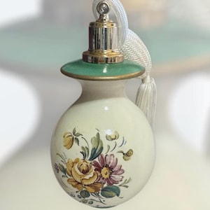 May include: A decorative perfume bottle with a cream-colored body and a floral design featuring yellow and purple flowers. The bottle has a green and gold top with a silver and gold metal cap and a white tassel.