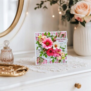 Pu&ograve; includere: Un biglietto d'auguri quadrato con sfondo a righe rosa e bianche, decorato con un bouquet di fiori di carta sovrapposti nei toni del rosa, bianco e verde. Il biglietto presenta una piccola etichetta bianca con il testo stampato Happy Birthday Christine.