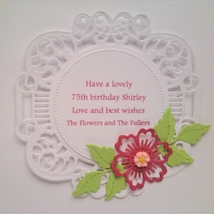 Pu&ograve; includere: Un biglietto circolare bianco con un intricato bordo in pizzo fustellato. Al centro &egrave; presente un testo rosa che recita: Have a lovely 75th birthday Shirley Love and best wishes The Flowers and The Fullers. Un fiore rosso con foglie verde chiaro.