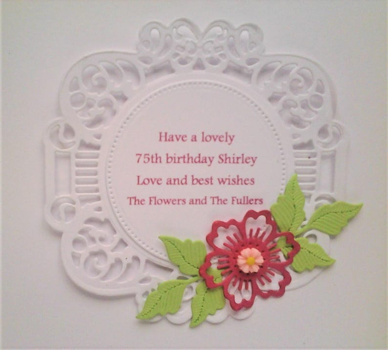 Pu&ograve; includere: Un biglietto circolare bianco con un intricato bordo in pizzo fustellato. Al centro &egrave; presente un testo rosa che recita: Have a lovely 75th birthday Shirley Love and best wishes The Flowers and The Fullers. Un fiore rosso con foglie verde chiaro.