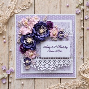 May include: A square greeting card with a light purple patterned background, decorated with layered dark purple and light pink fabric flowers, white beaded accents, and a white die-cut lace border. The text reads Happy 85th Birthday Auntie Beth.