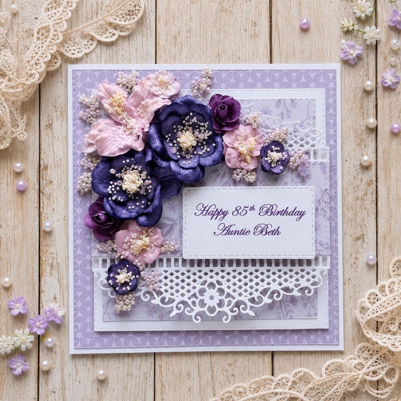 May include: A square greeting card with a light purple patterned background, decorated with layered dark purple and light pink fabric flowers, white beaded accents, and a white die-cut lace border. The text reads Happy 85th Birthday Auntie Beth.