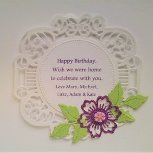 May include: A white circular card with an intricate die-cut lace border. The centre features the purple text: Happy Birthday. Wish we were home to celebrate with you. Love Mary, Michael, Luke, Adam & Kate. A purple glitter flower with green leaves.