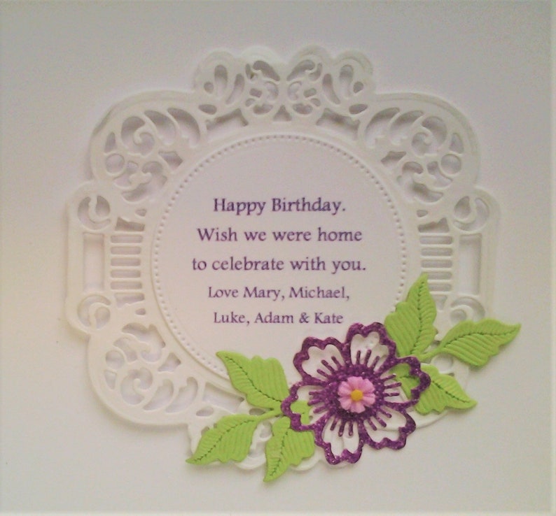 May include: A white circular card with an intricate die-cut lace border. The centre features the purple text: Happy Birthday. Wish we were home to celebrate with you. Love Mary, Michael, Luke, Adam & Kate. A purple glitter flower with green leaves.